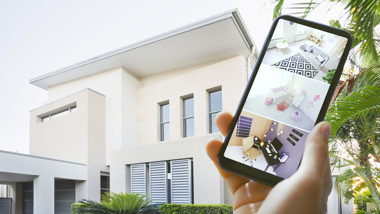 A hand holding a smartphone showing live security camera feeds in front of a contemporary home.